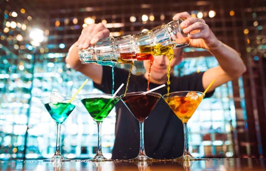 Bartender pours colorful alcoholic cocktails in martini glasses at the bar.