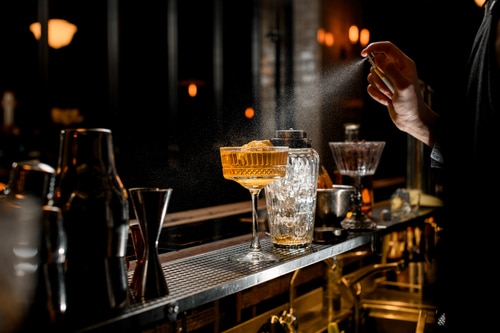 Professional bartender in black apron energetically sprinkles on glass with cold brown alcoholic cocktail on bar counter after reviewing the Ultimate Guide to bartending. Blurred view of restaurant in background.