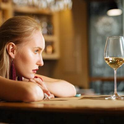 A blonde woman leaning her head on her arms on a bar table next to a glass of white wine.