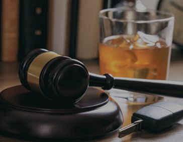 A judge's gavel and car keys on a desk with a glass of amber liquor in the background.
