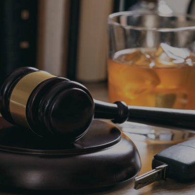 A judge's gavel and car keys on a desk with a glass of amber liquor in the background.
