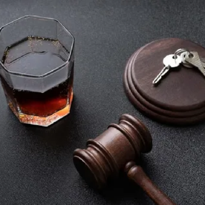 A judge's gavel and car keys on a desk with a glass of amber liquor in the background.
