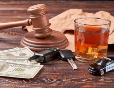A gavel, car keys, cash, a drink, and a toy police car on a wooden table.