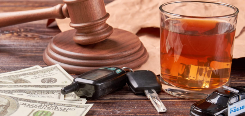 A gavel, car keys, cash, a drink, and a toy police car on a wooden table.