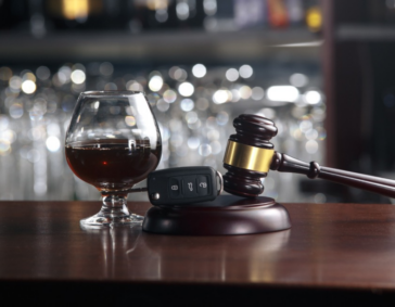 A snifter of dark liquor sitting next to a car key fob and a wooden judge's gavel on a bar top.