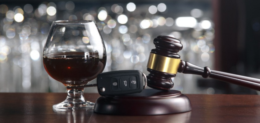 A snifter of dark liquor sitting next to a car key fob and a wooden judge's gavel on a bar top.