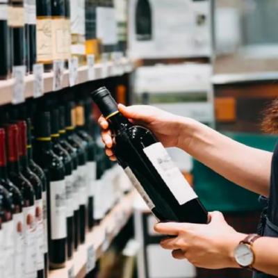 A customer or employee in a retail store holding a bottle of wine while browsing a well-stocked shelf of red wine.